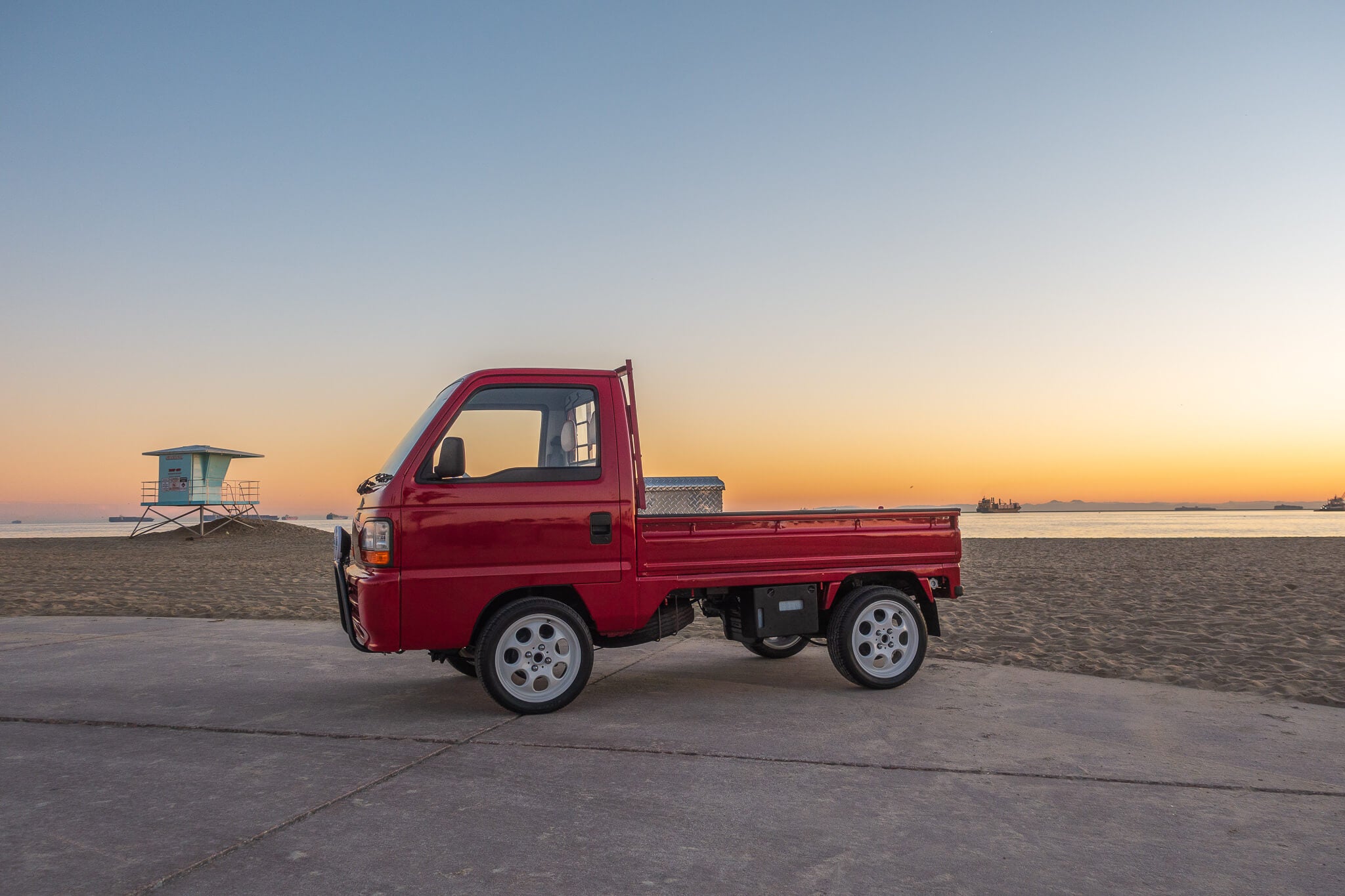 Red Honda Acty Kei Truck Parked on a Beach at Sunset with a Lifeguard Tower in the Background