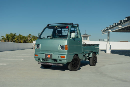 Mint green Honda kei truck parked in an outdoor setting with clear blue sky and palm trees in the background, showcasing the vehicle's compact design and sturdy build.