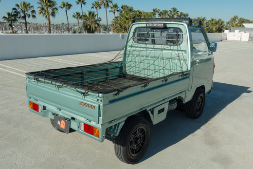 Rear view of a mint green Honda kei truck with a netted cargo bed, parked in a sunny outdoor setting with palm trees in the background.