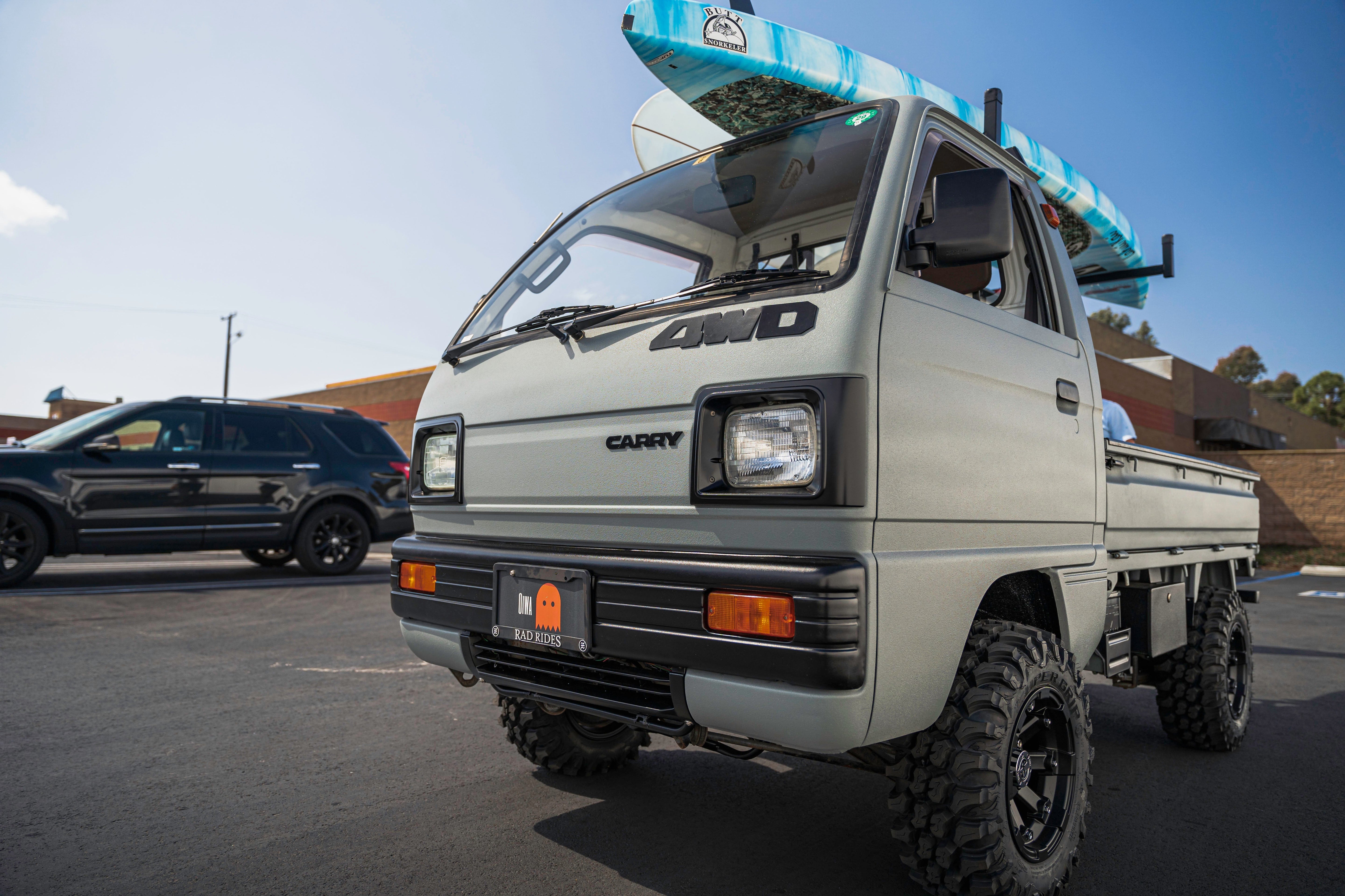 A small 4WD truck with a surfboard on top is parked in a lot, with another vehicle in the background.