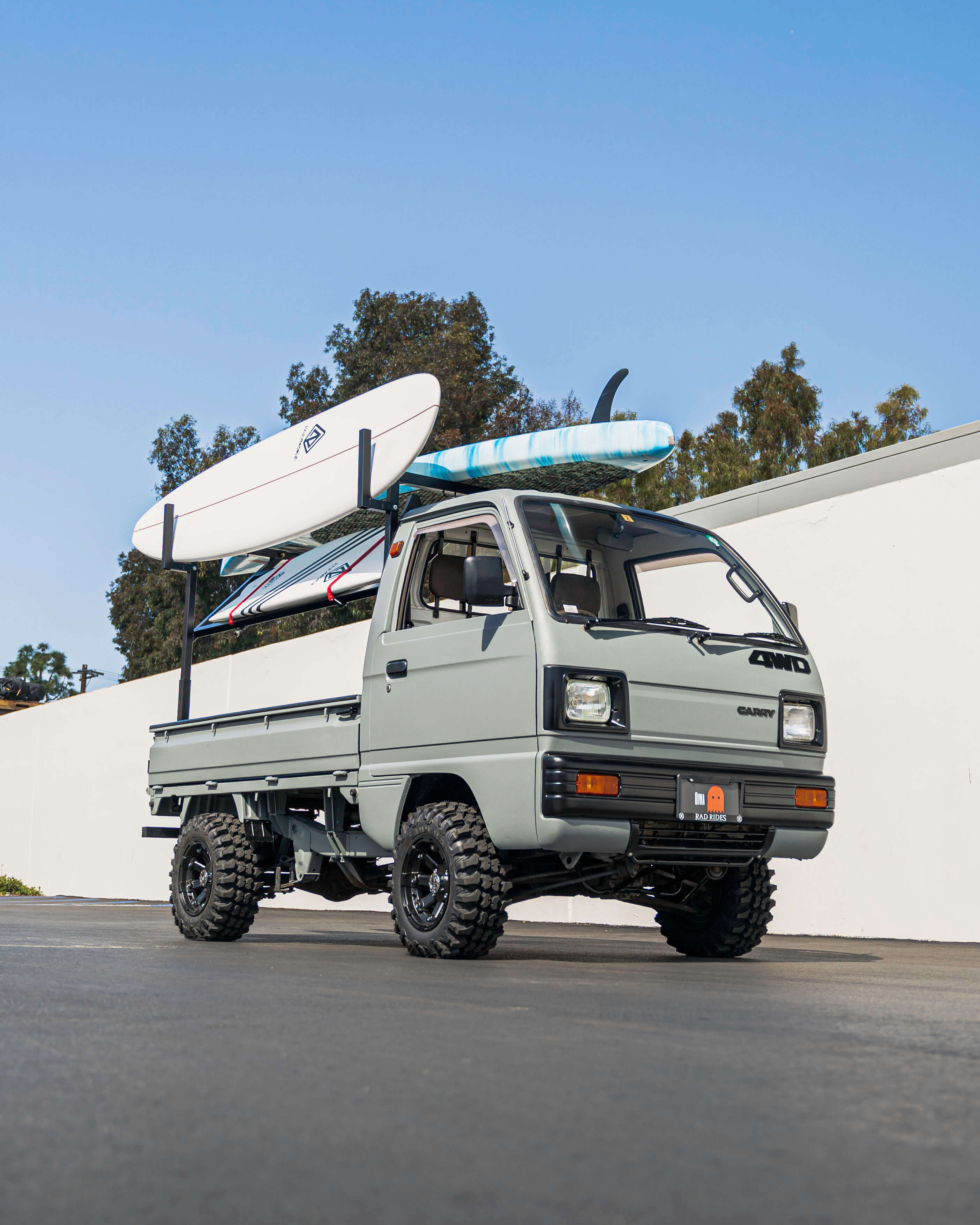 4WD Suzuki Carry Kei Truck with Surfboards Mounted on Roof Rack in a Sunny Outdoor Setting