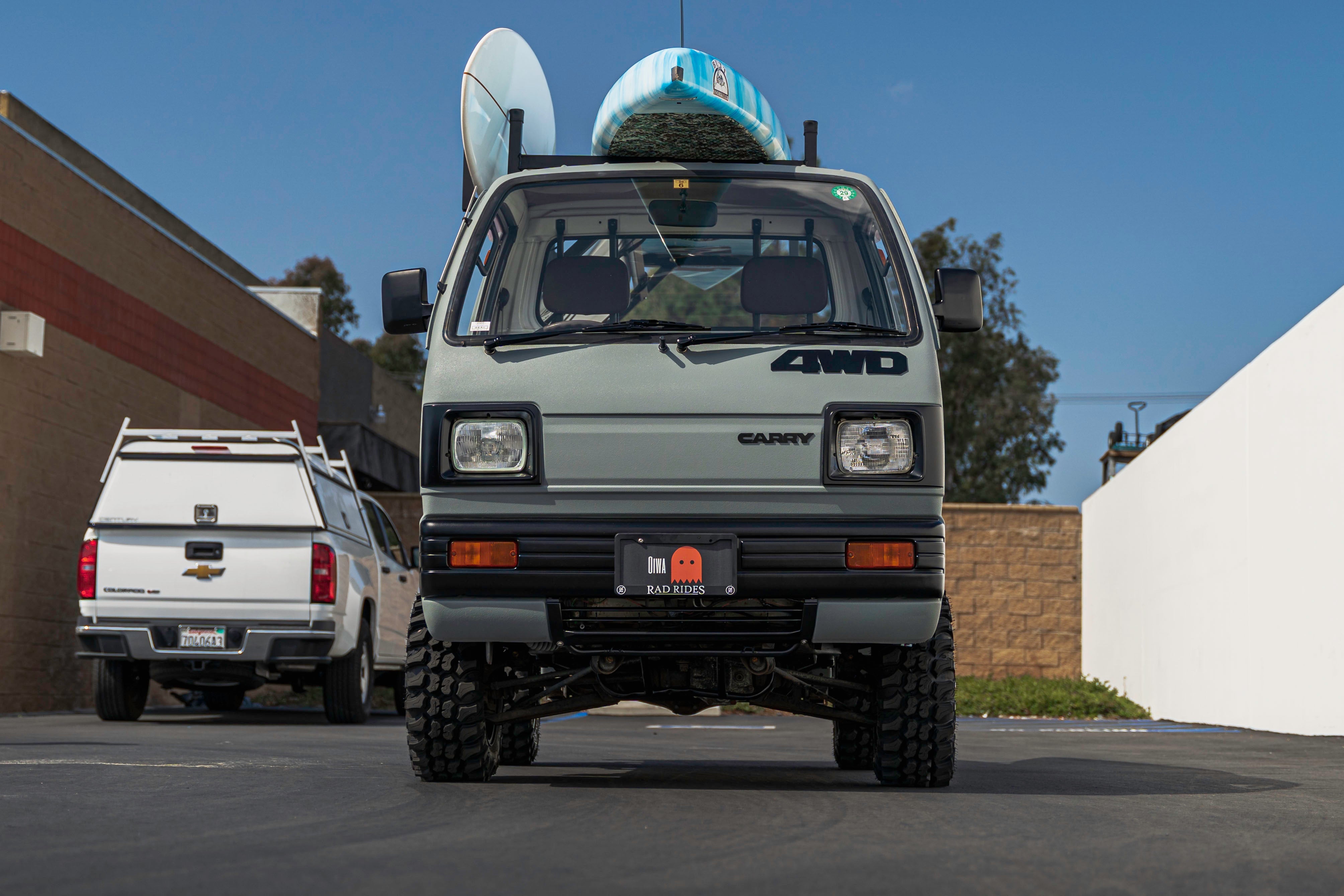 A front view of a 4WD mini truck with surfboards on the roof, parked in a lot next to a white pickup truck.