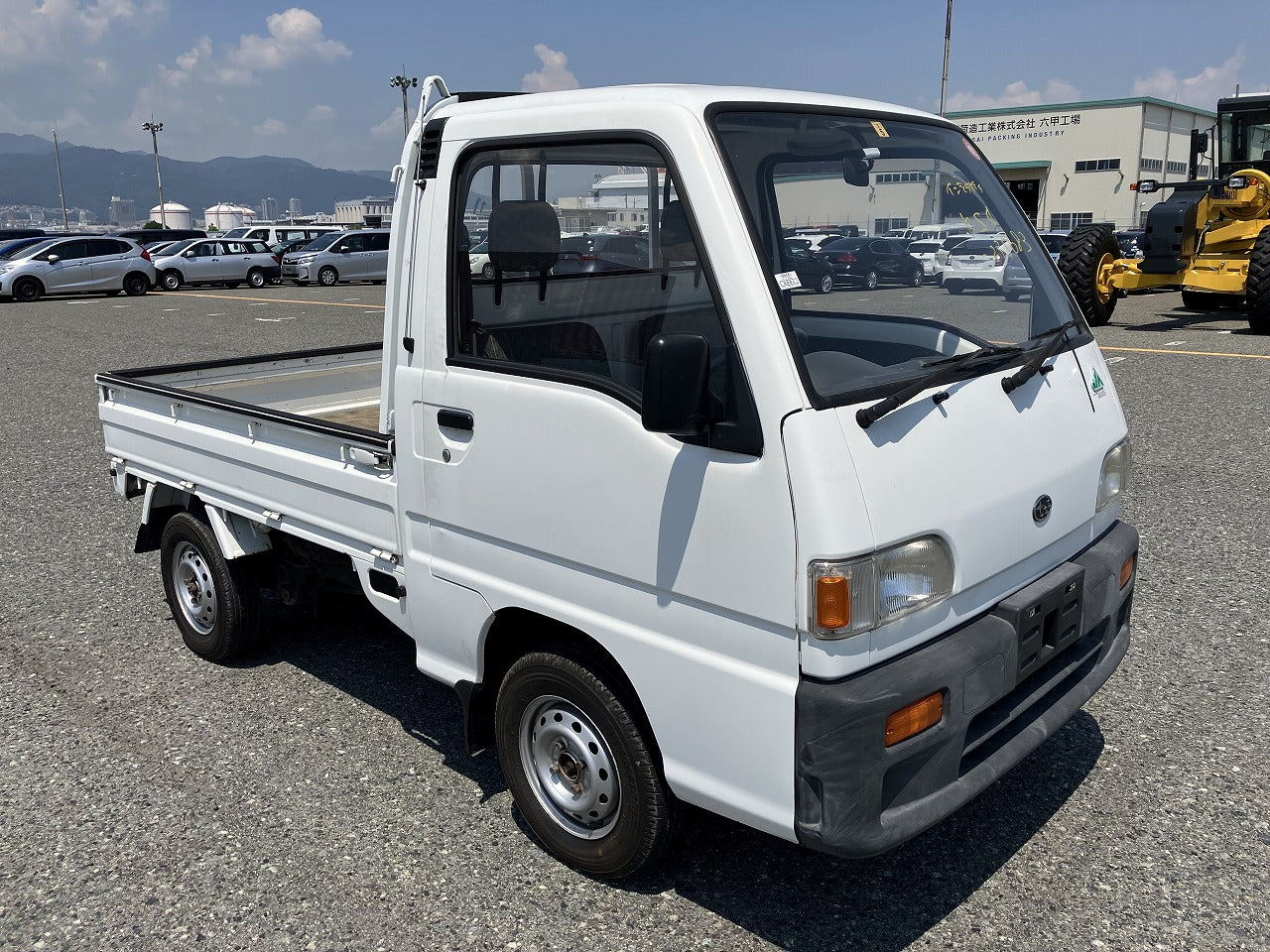 A small white pickup truck is parked in a lot with other vehicles and industrial equipment in the background.