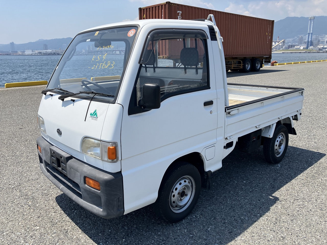 A small white pickup truck parked near a waterfront with shipping containers in the background.