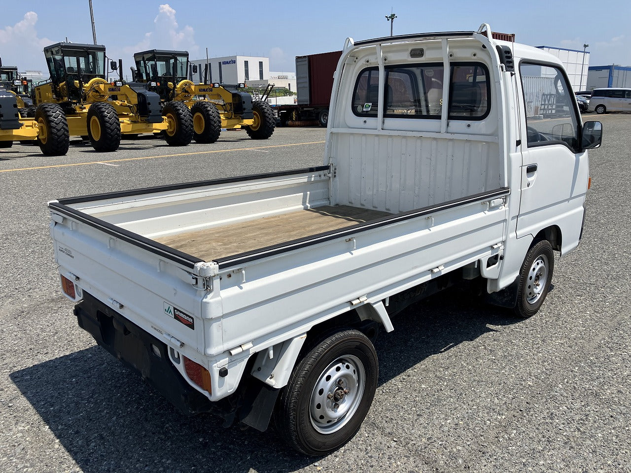 A small white pickup truck with an open cargo bed is parked on a paved lot, with several yellow construction vehicles in the background.