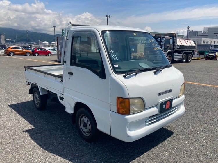 Front-side view of a Subaru Sambar kei truck parked at a lot in Seattle, showcasing its compact design, 4WD capability, and practicality. Perfect for city driving or light-duty tasks, this Japanese import is available for sale through Oiwa Garage.