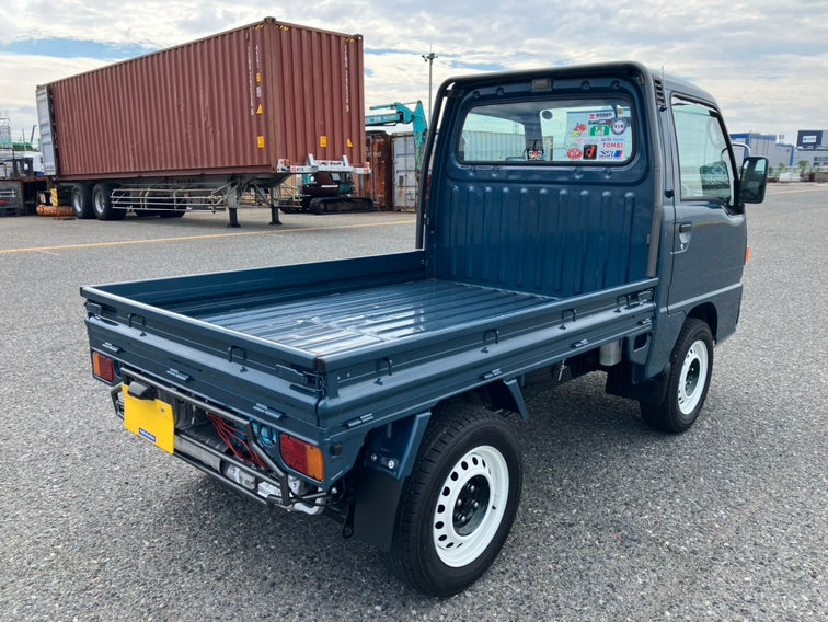 Subaru Sambar kei truck parked by a waterfront with scenic mountain view.