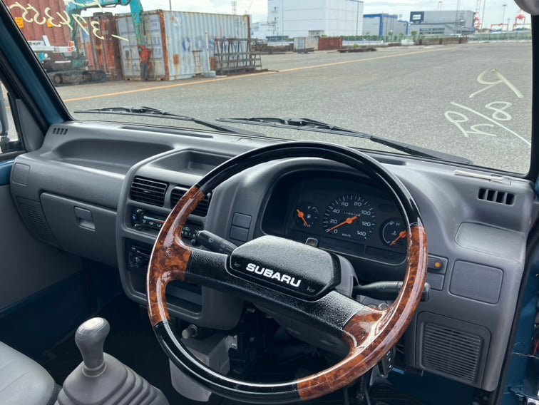 Interior view of a Subaru Sambar showing steering wheel, dashboard, and manual gear shift with industrial area visible.