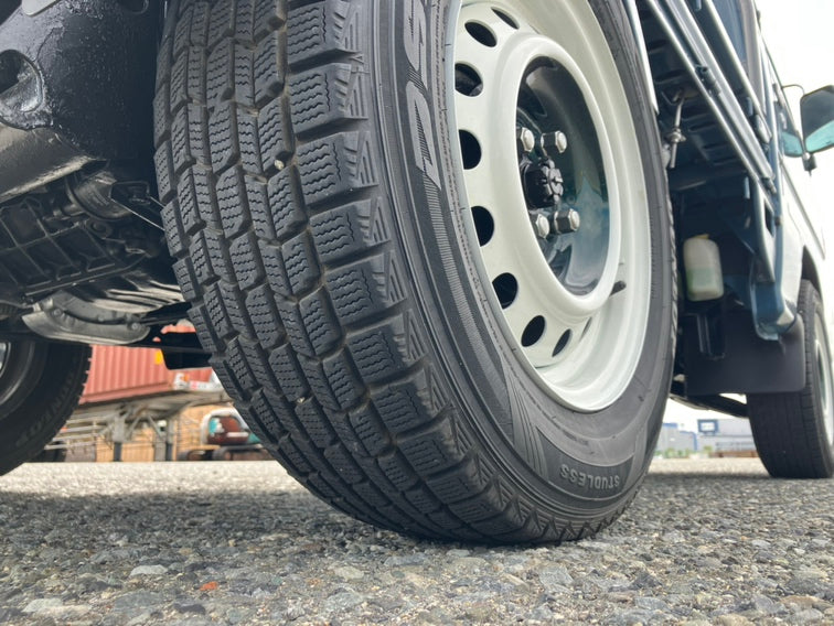 Close-up of a 1969 Subaru Sambar tire with detailed tread, white wheel on gravel, red container in background, showcasing the kei truck's rugged charm.