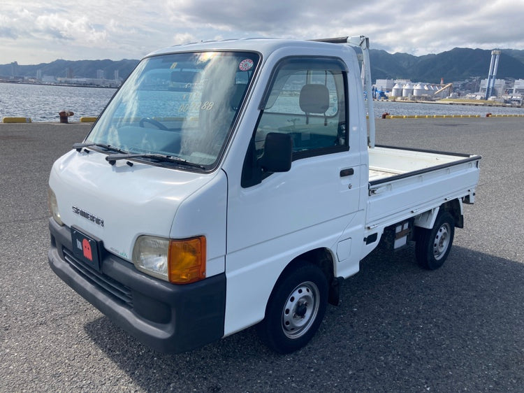 White Subaru Sambar kei truck parked near the waterfront, showcasing its compact size and versatility. Ideal for import to Florida with Oiwa.co and parts from OiwaGarage.co.