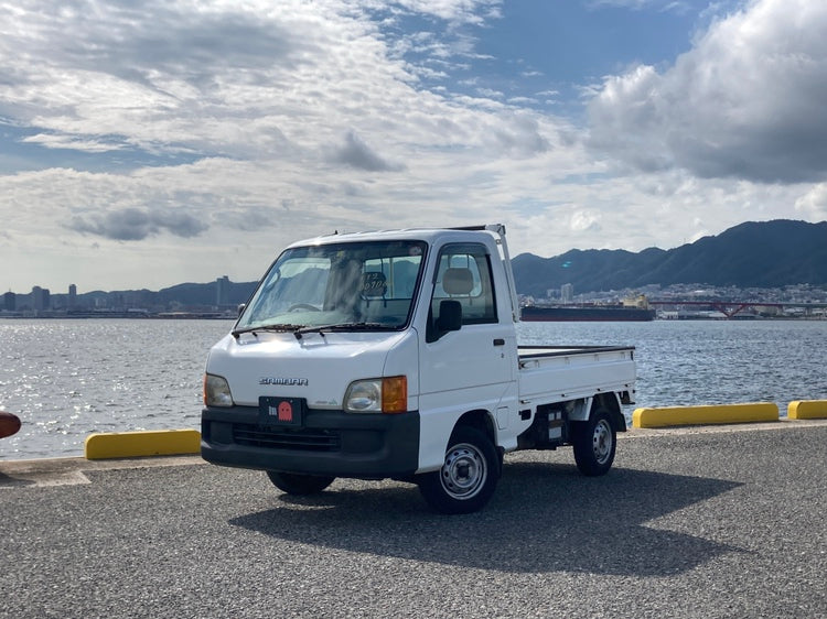 White Subaru Sambar kei truck parked by a scenic waterfront, showcasing its compact design and versatility, perfect for urban and rural use. Import yours with Oiwa.co.