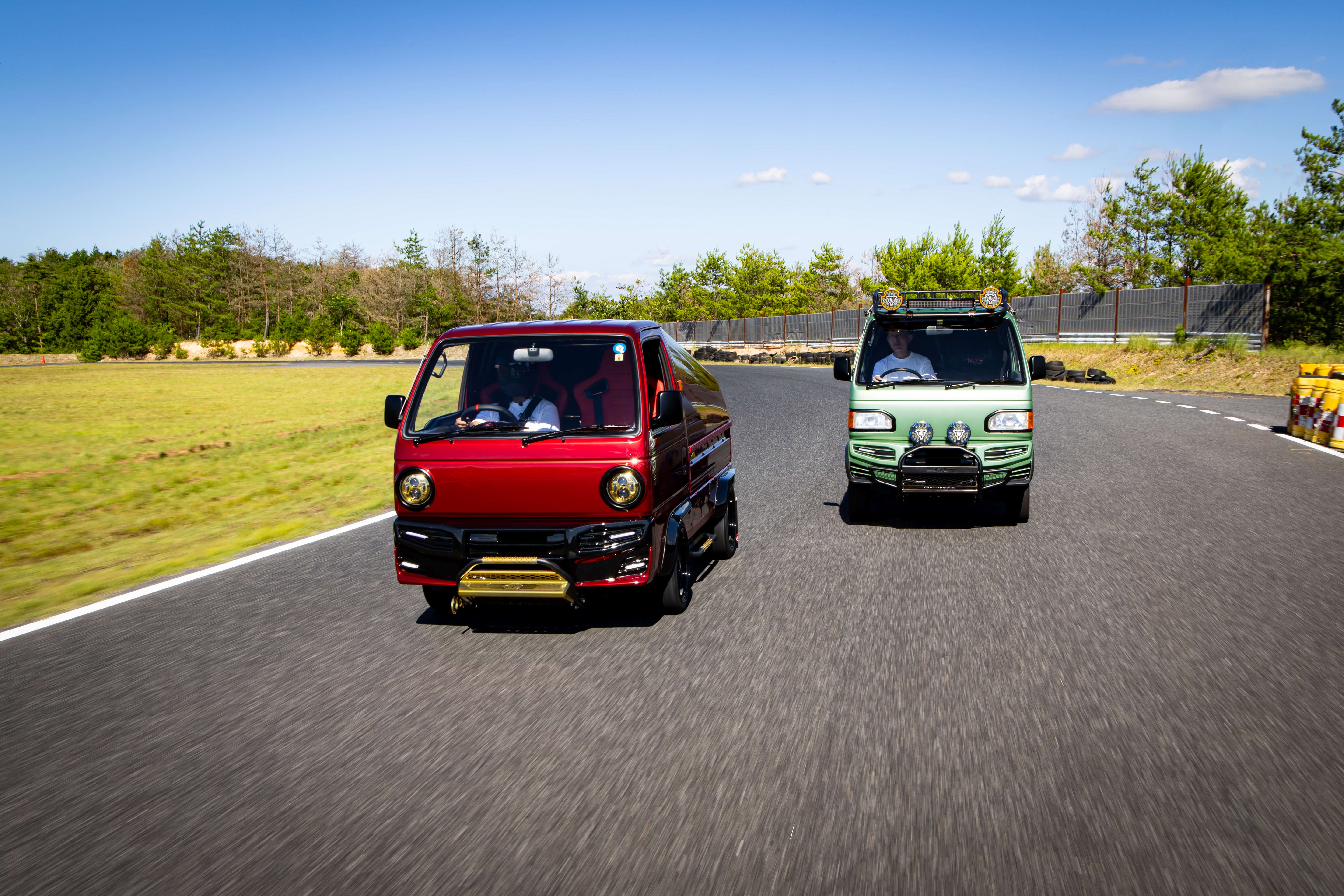 A red, performance-tuned Honda Acty kei truck races side-by-side with a green, off-road-styled Honda Acty on a test track, each showcasing their impressive top speeds against a scenic backdrop of greenery.