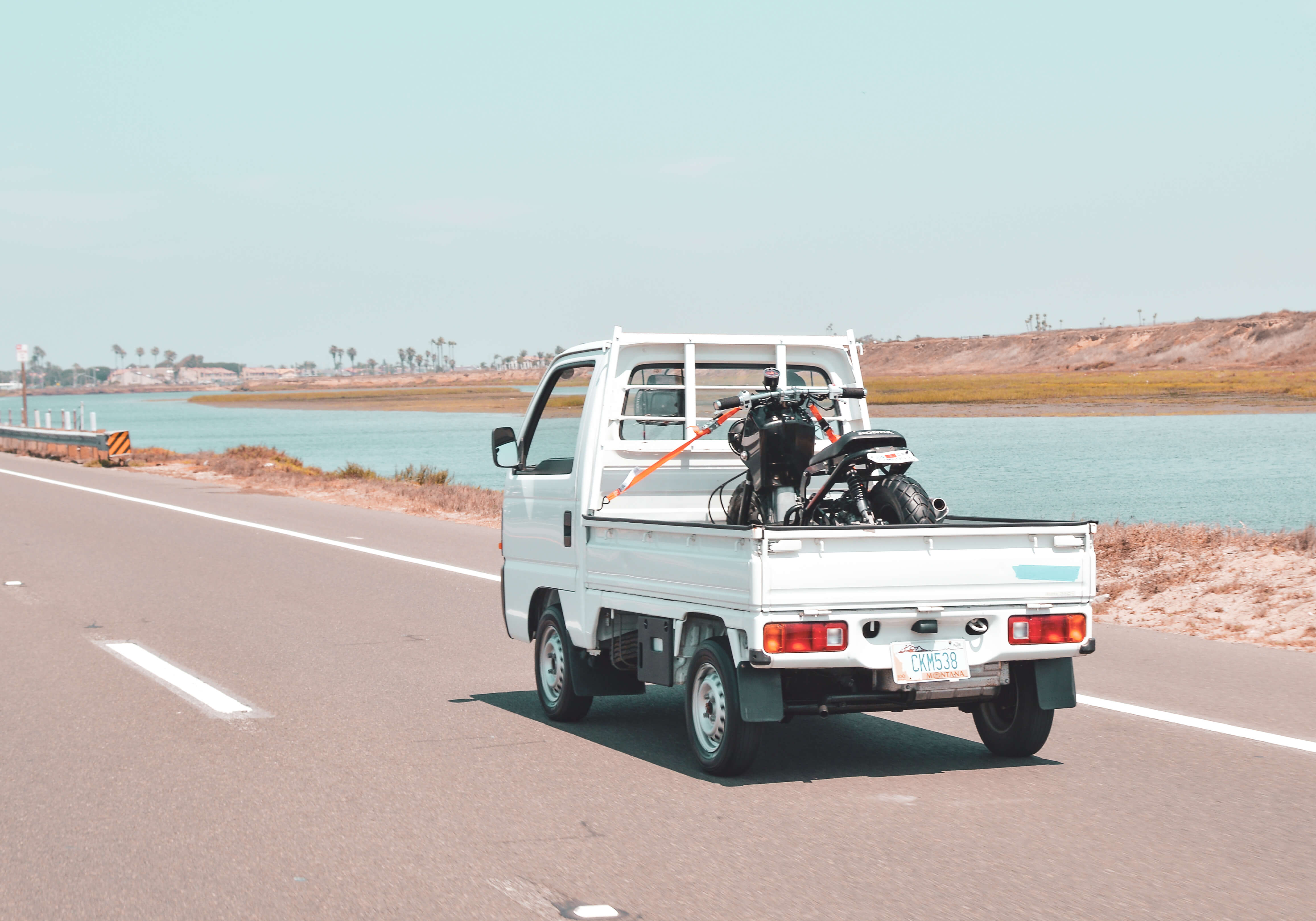 White Honda Acty mini truck carrying a motorcycle on an open road by a scenic waterway, showcasing its versatility and fuel efficiency on a long-distance journey from York, Maine to Miami, Florida.