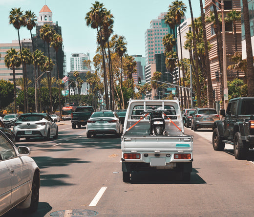 Vintage white Honda Acty truck carrying cargo on a busy downtown street, surrounded by modern cars and palm trees, showcasing urban transportation diversity.