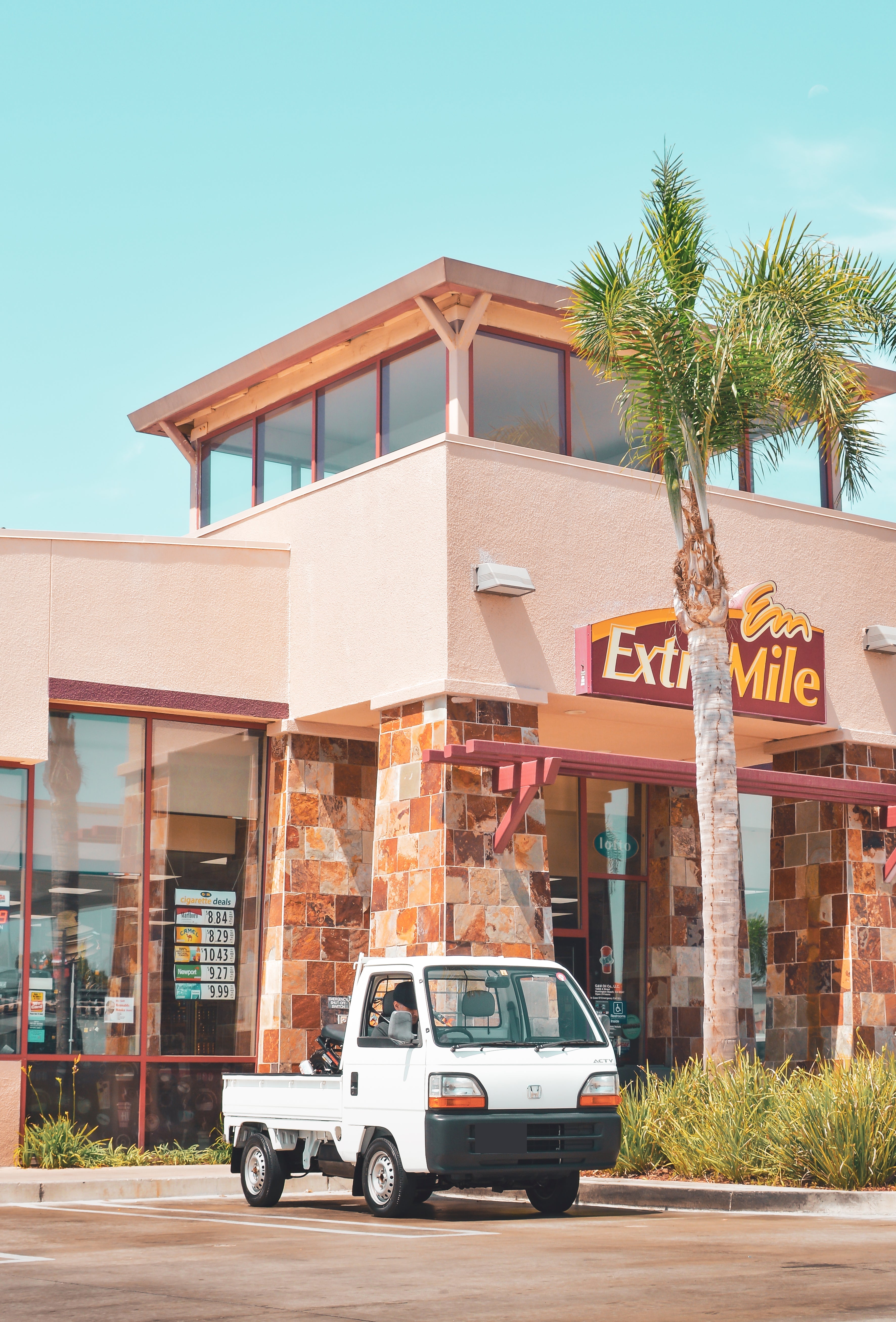 White Honda Acty mini truck parked outside ExtraMile convenience store in a sunny suburban setting, showcasing compact size and urban functionality.