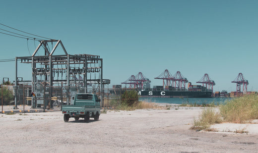 A mint green kei truck driving on a gravel path near industrial power lines and shipping cranes with a large cargo ship in the background under a clear blue sky.