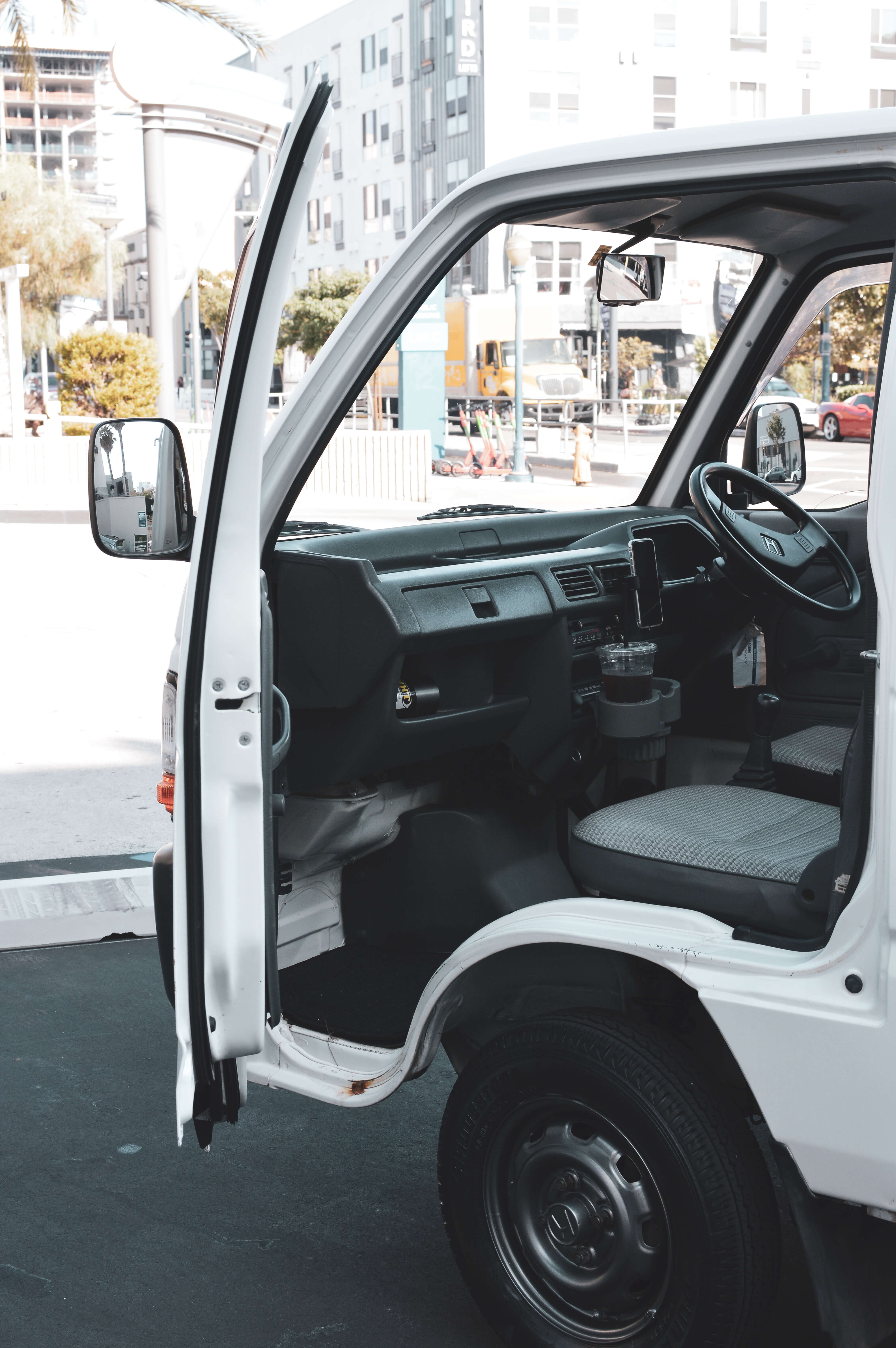 Open driver's side door of a white 4 door kei truck, revealing gray and black interior, on a city street with trees and a yellow truck in the background.