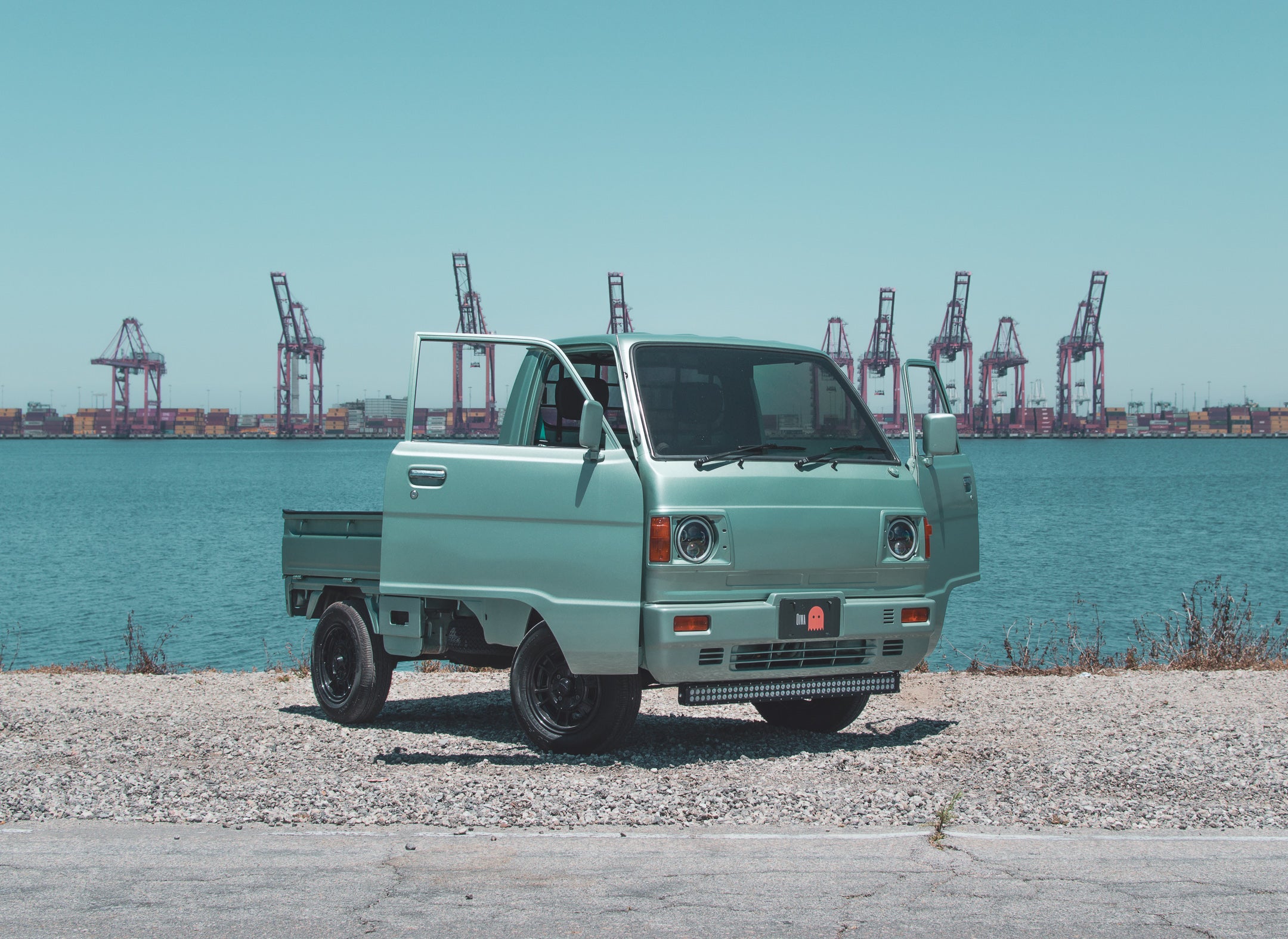 Modern kei truck parked on gravel near water with cranes and containers in the background, showcasing compact and versatile design.