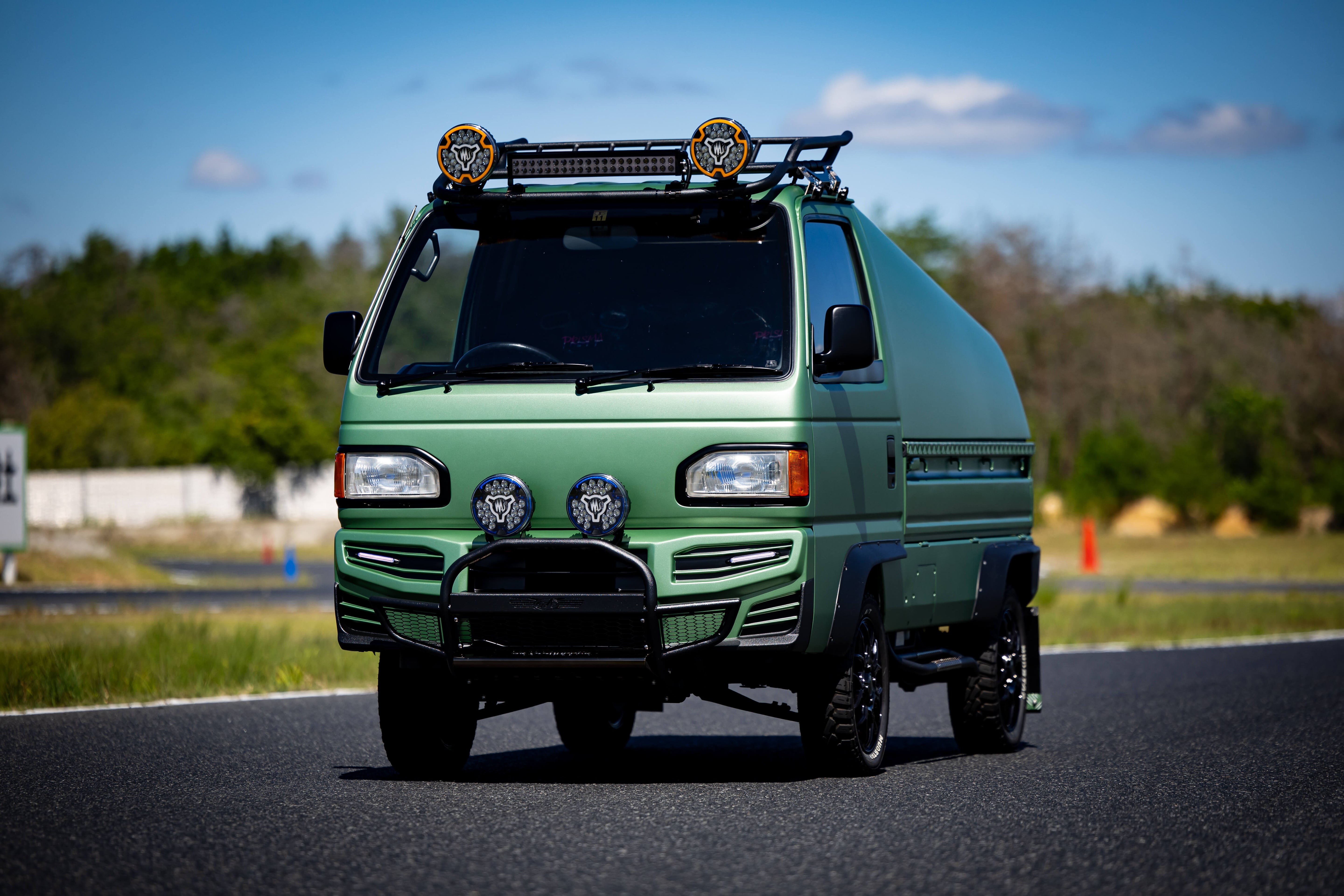 A green offroad kei truck with roof rack and lights parked on a paved road, set against a backdrop of trees and clear blue sky.