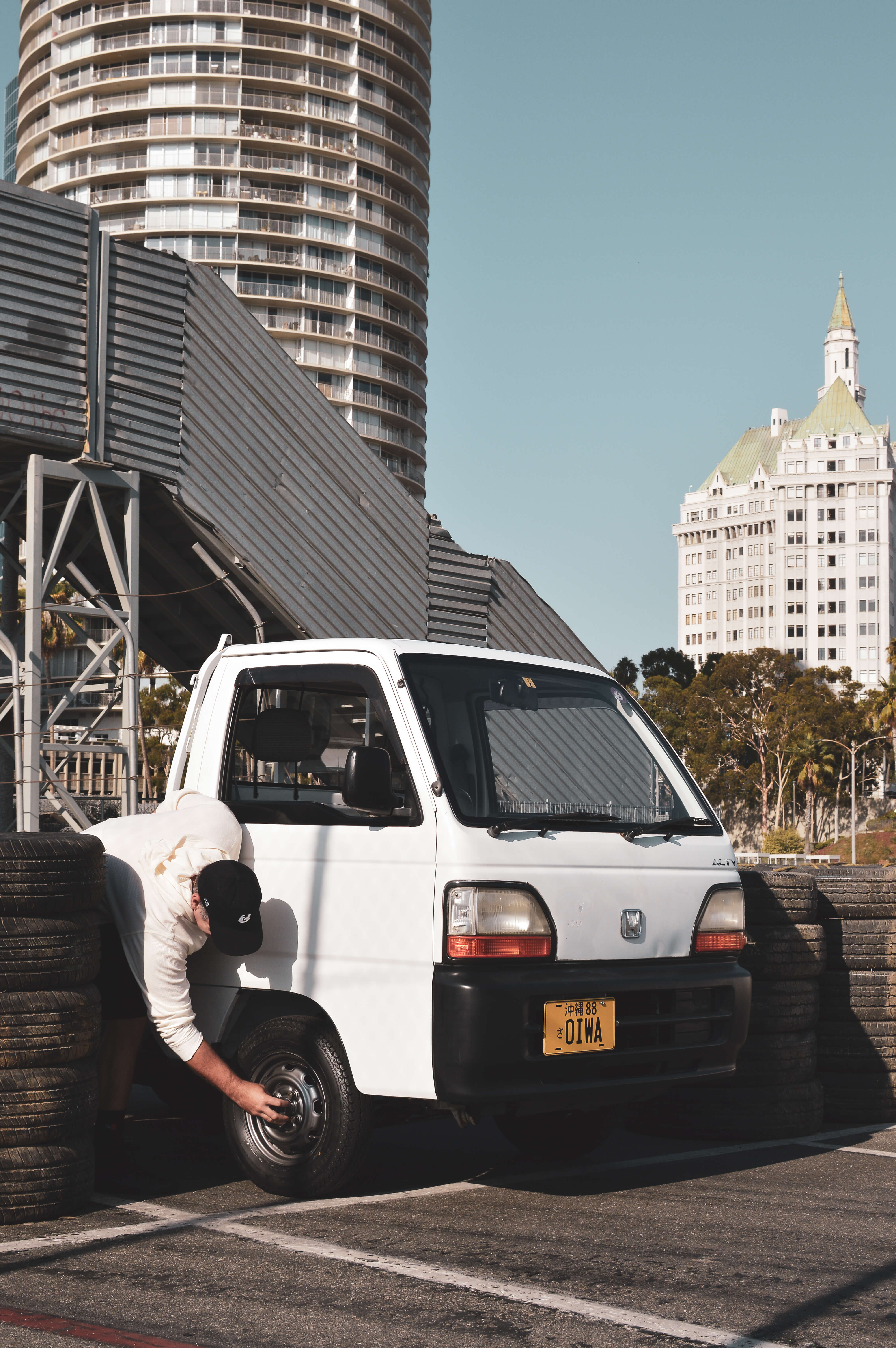 A person examining the front wheel of a JDM kei truck with yellow plates, surrounded by tall buildings and tires.