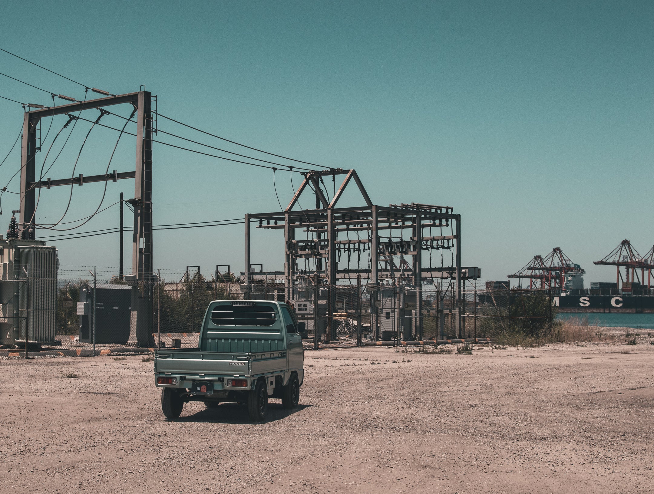 A custom green kei truck parked in an industrial area with cranes and a cargo ship, symbolizing Japanese automotive innovation.