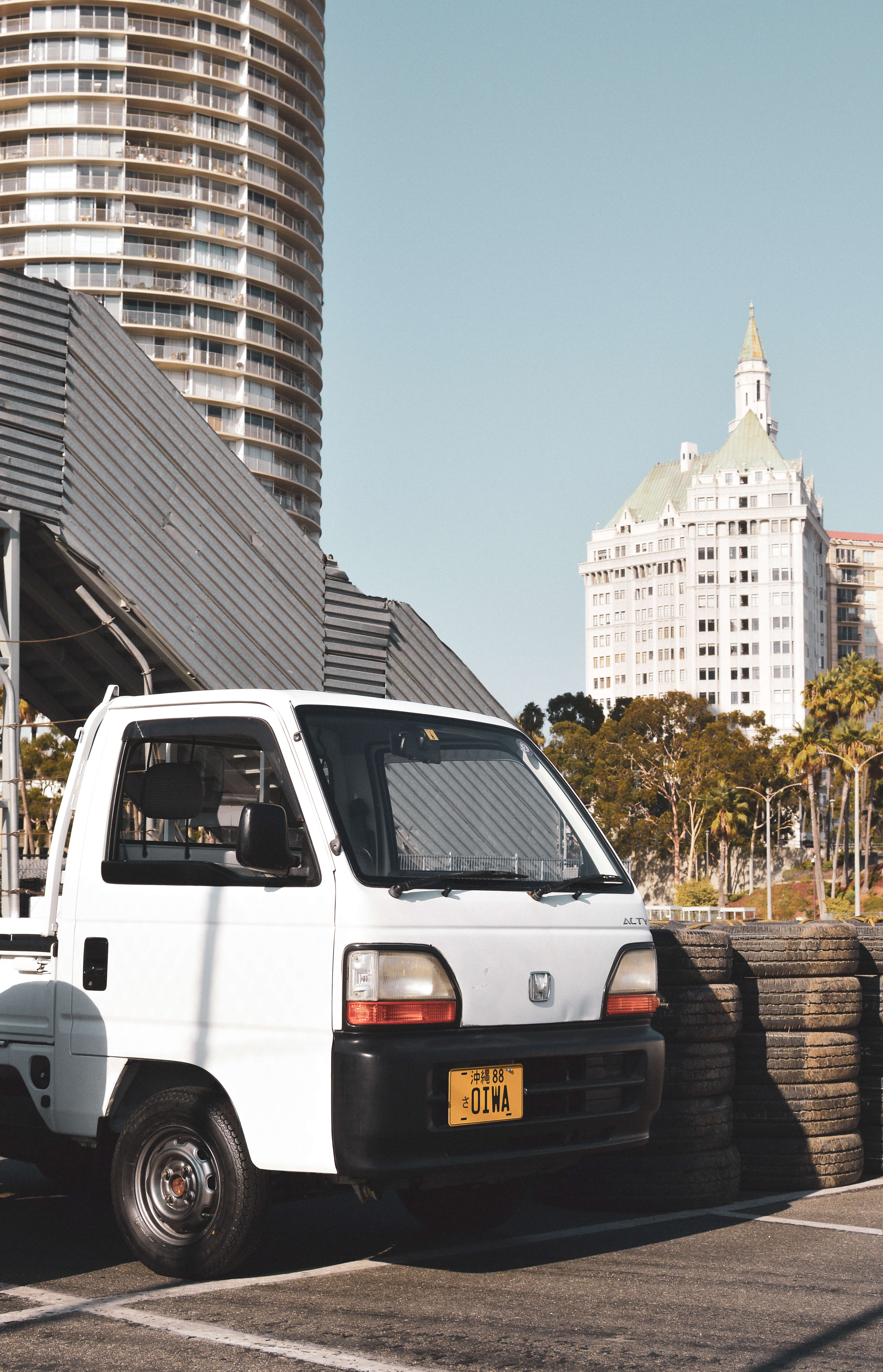 Lifted white kei truck parked beside tires with cityscape background, showcasing miniature truck modifications.