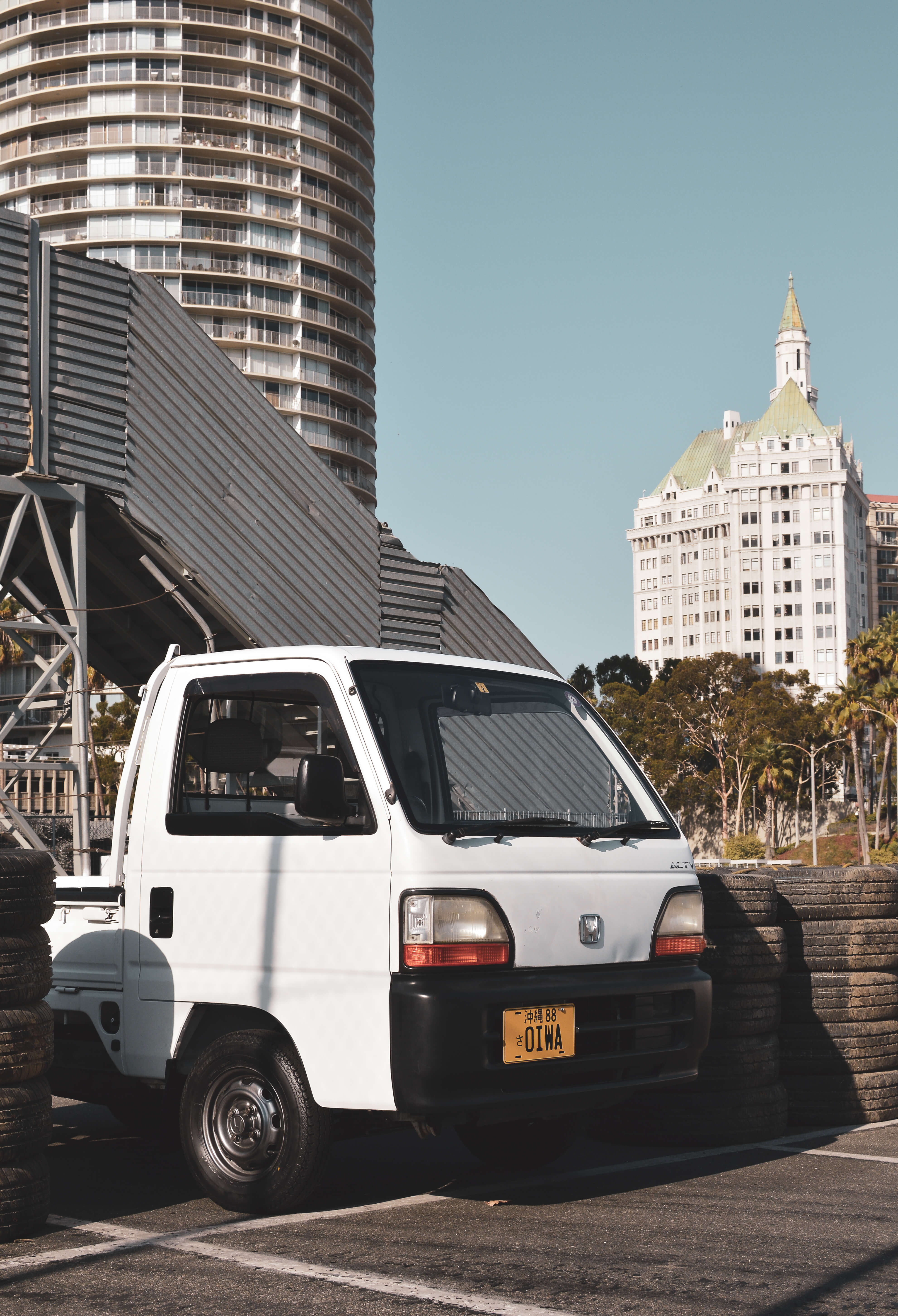 Import kei truck parked in front of tires, with buildings and trees in the background, under a clear blue sky.