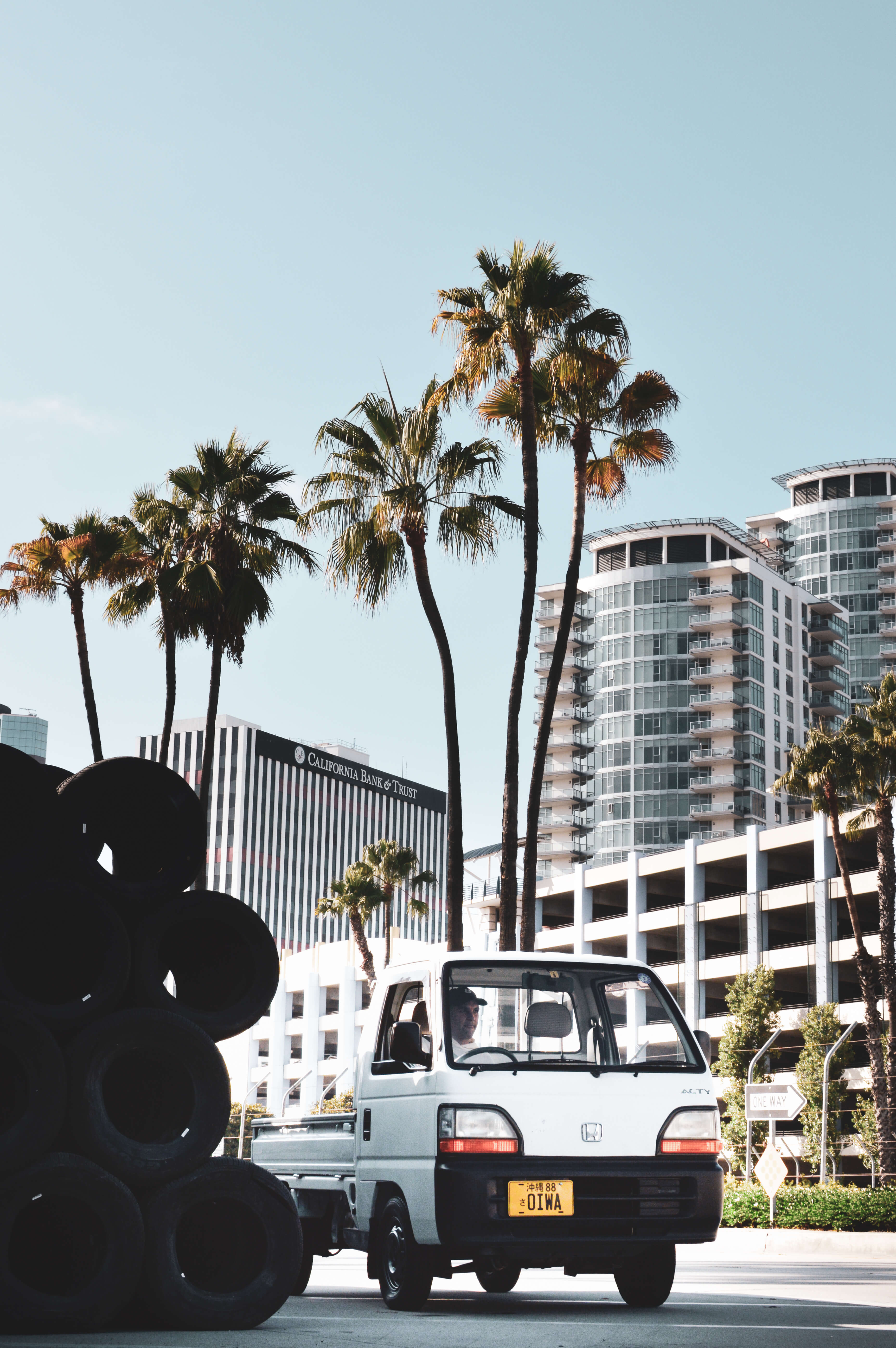 A white Japan kei truck parked on a street with tall palm trees and modern buildings in the background under a clear sky.