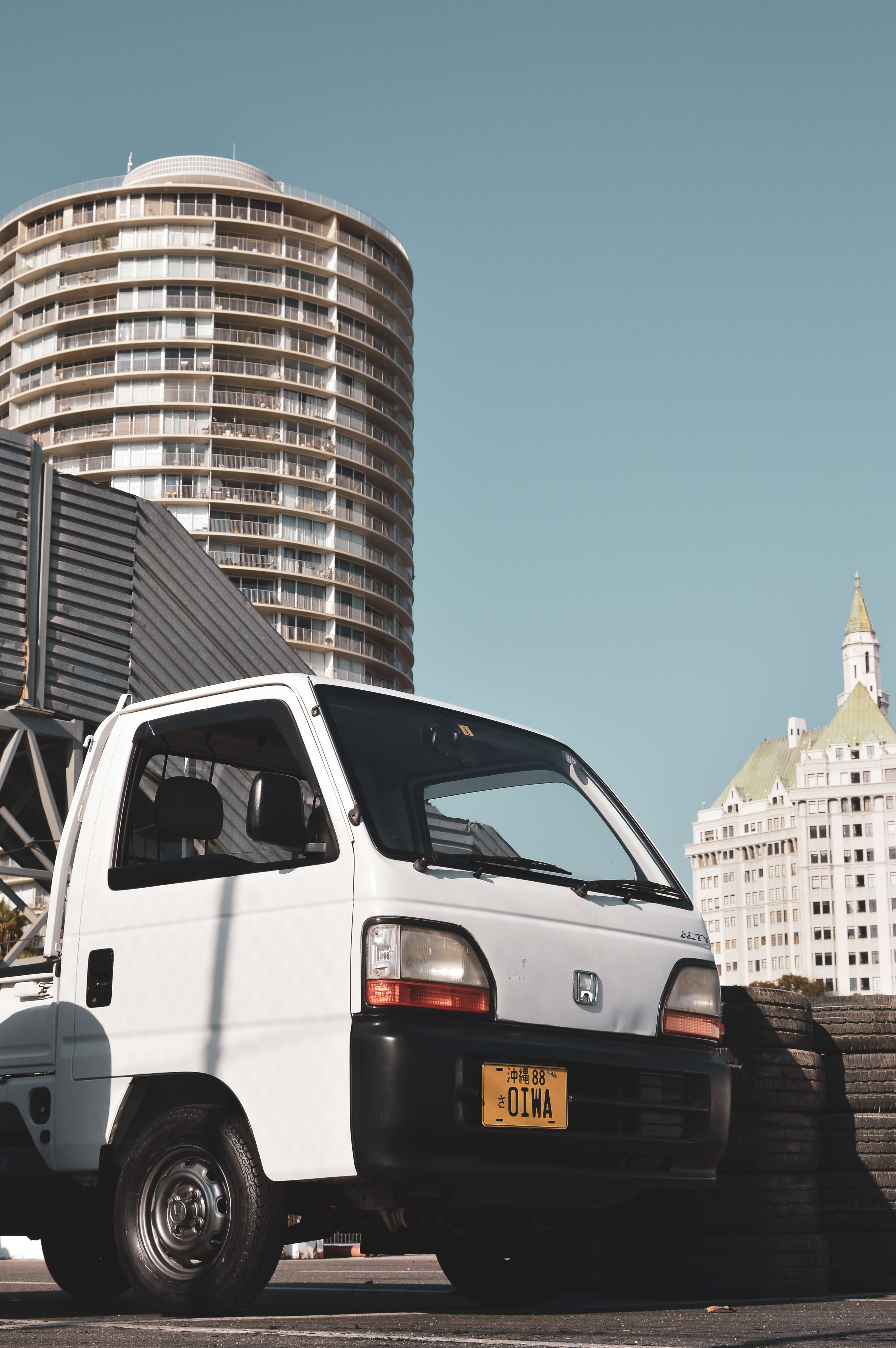 White kei truck with yellow license plate parked by tires, tall building in background, clear blue sky.