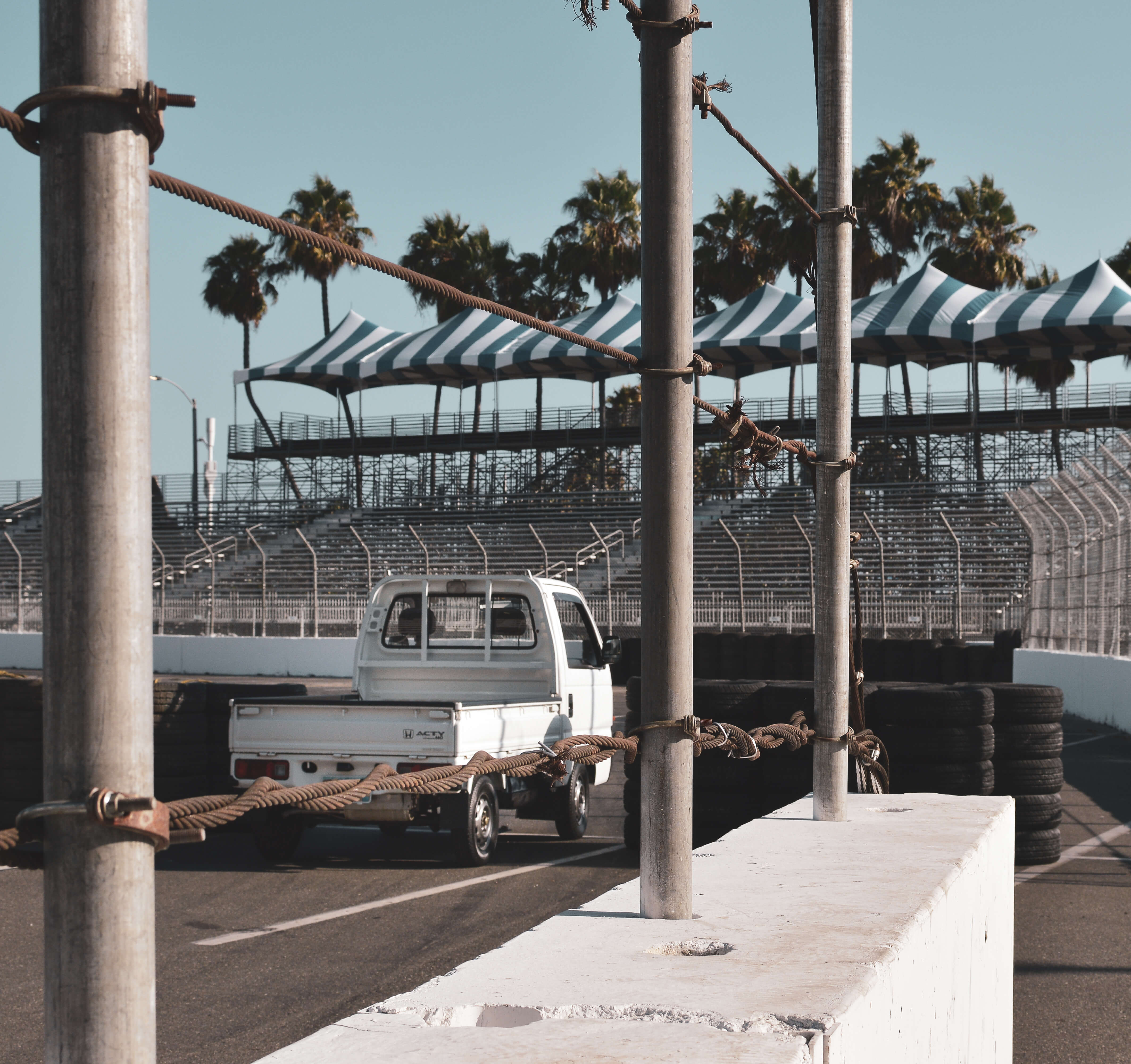 White kei truck parked on racetrack with grandstands and palm trees under clear blue sky.