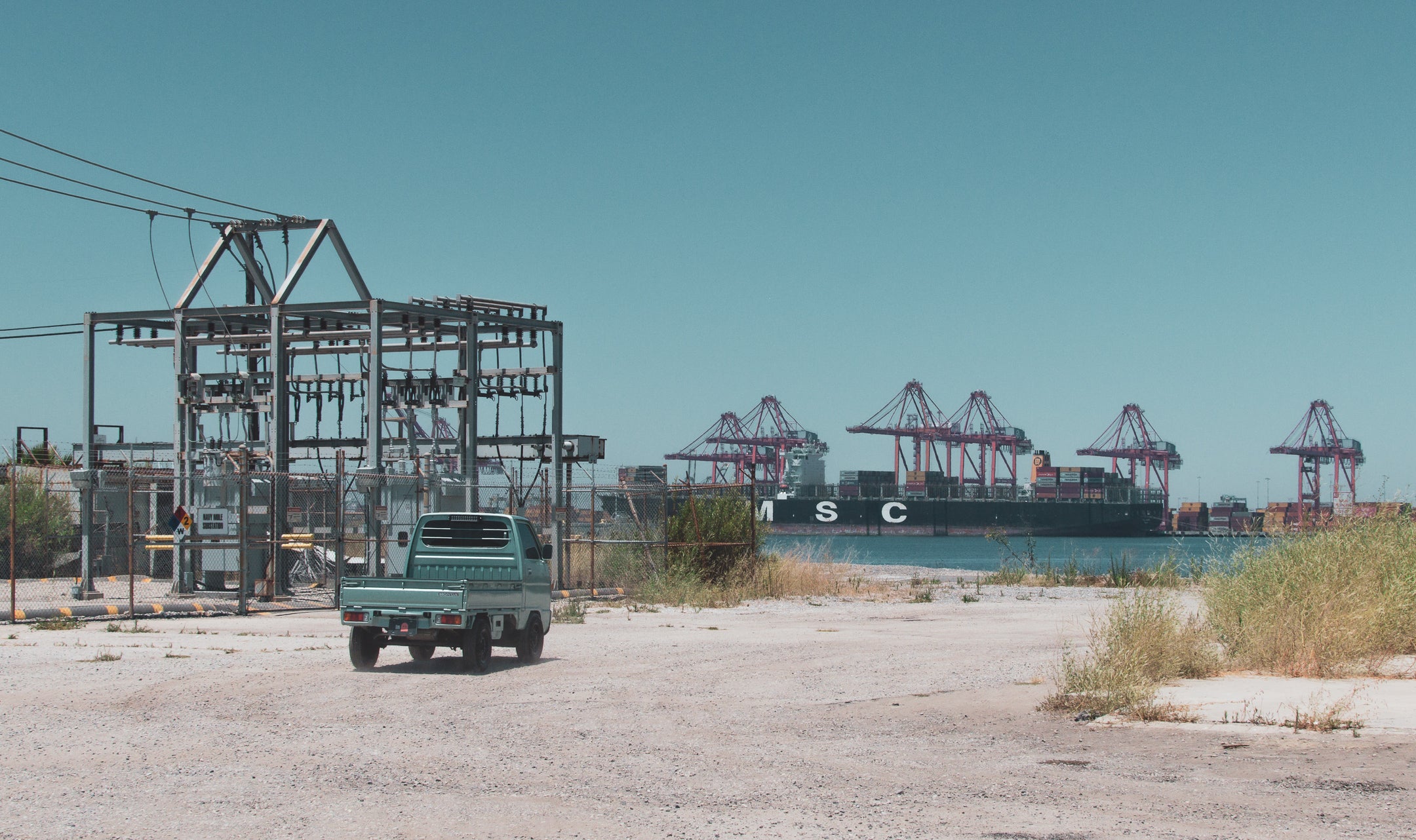 Light blue kei truck parked near industrial structures and cargo ship, showcasing potential for accessories.