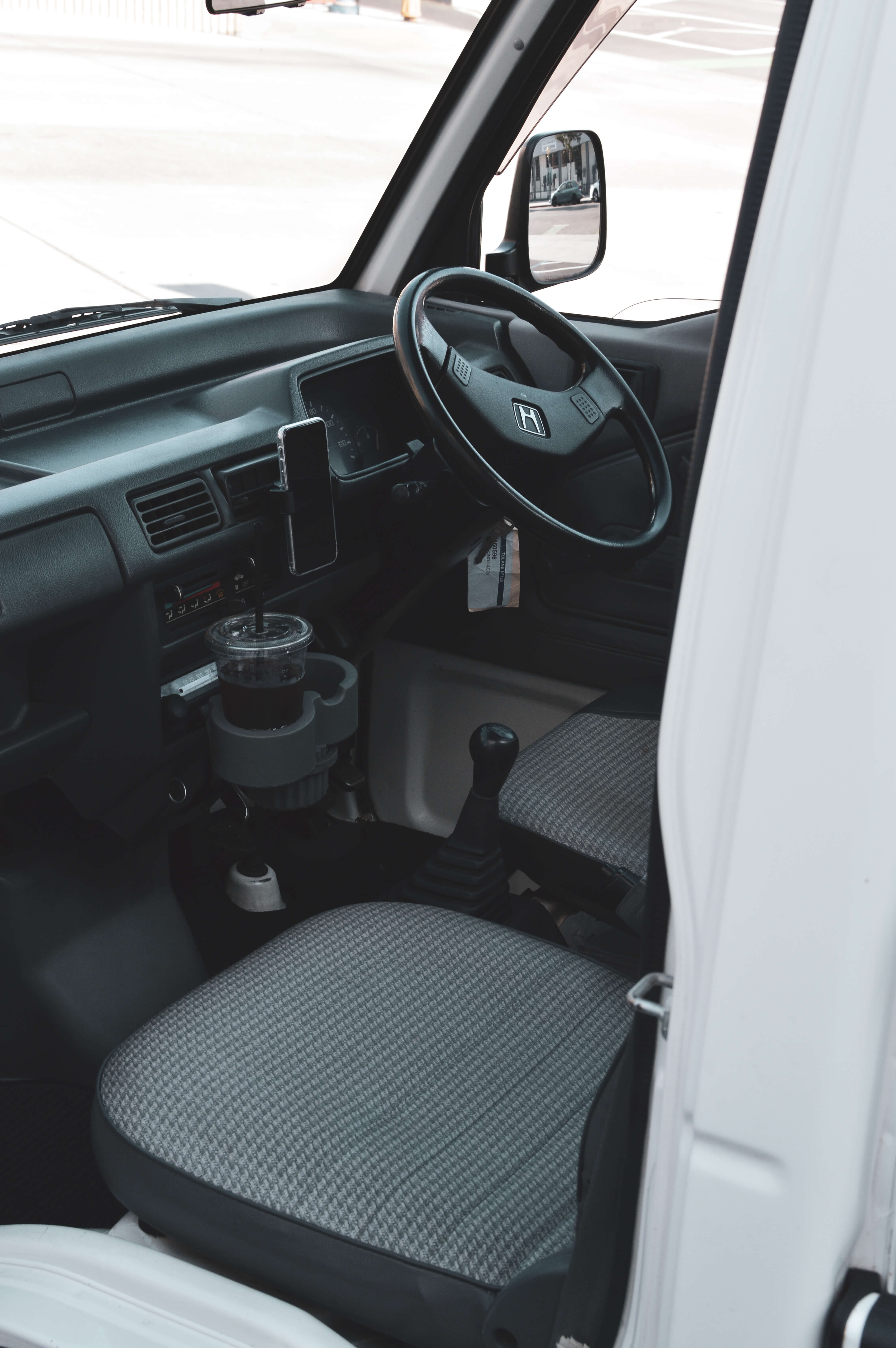 Interior view of a Nissan Kei Truck focusing on the dashboard, smartphone holder, and cup holder. The steering wheel has a logo, and gray fabric seats are visible.