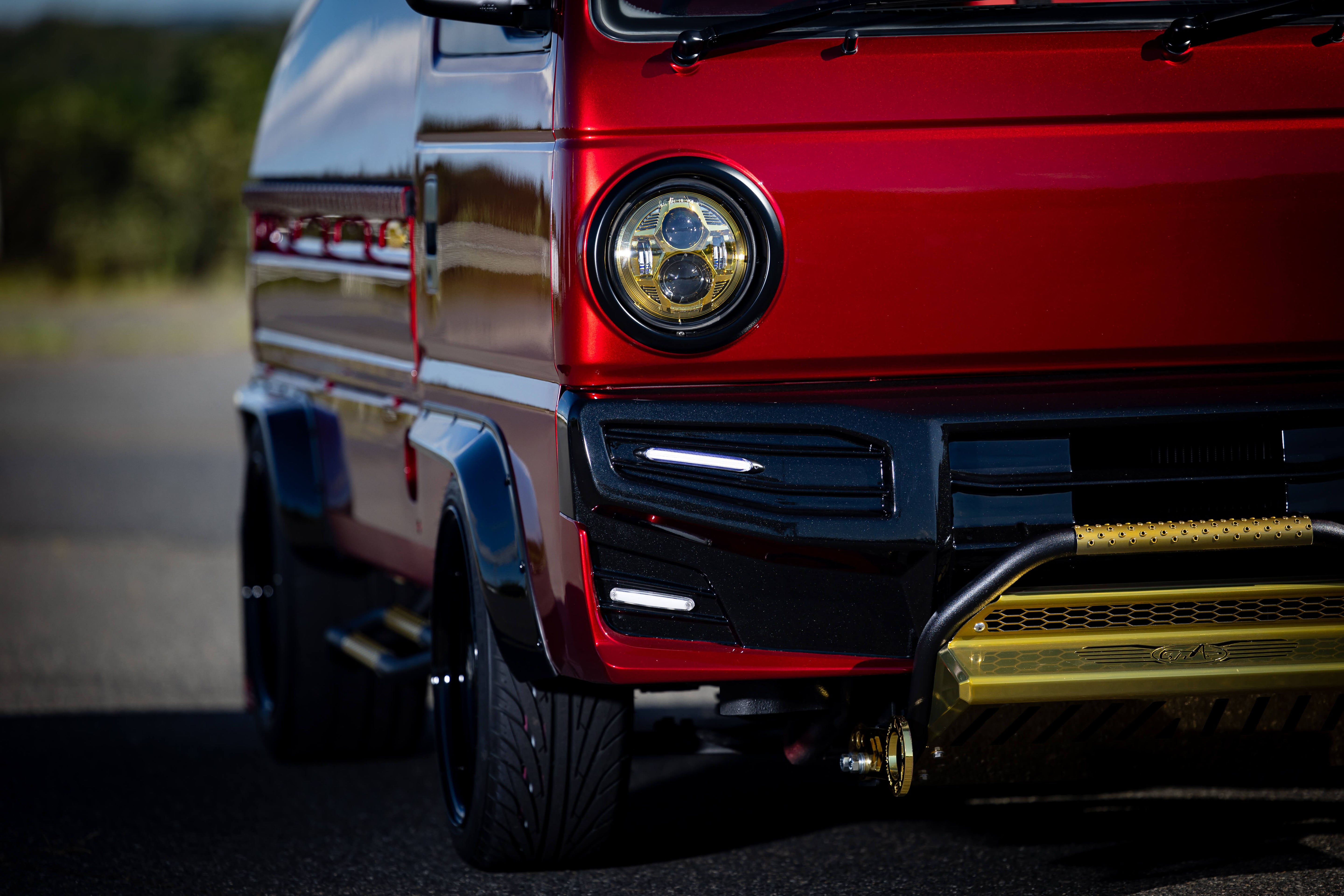 Oiwa kei truck with glossy red exterior, black accents, round headlight, and gold grille, on a road with a blurred natural background.