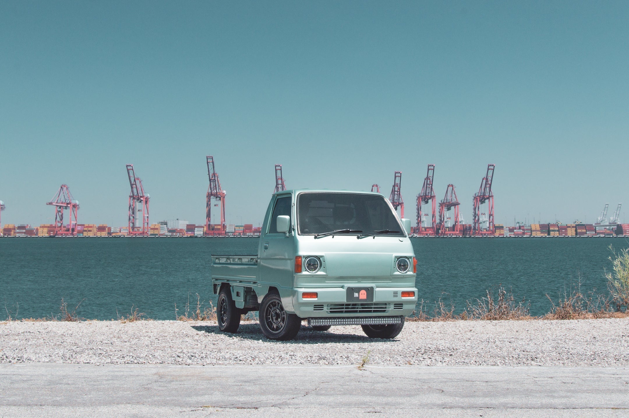 Subaru Sambar kei truck parked at a port with cranes and containers, showcasing its compact design and utility.