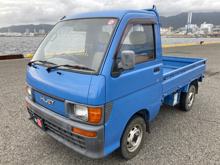 Blue 1997 Daihatsu Hijet S110P mini truck showcasing clean exterior, original headlights, and windshield markings, parked near waterfront with industrial mountain landscape.