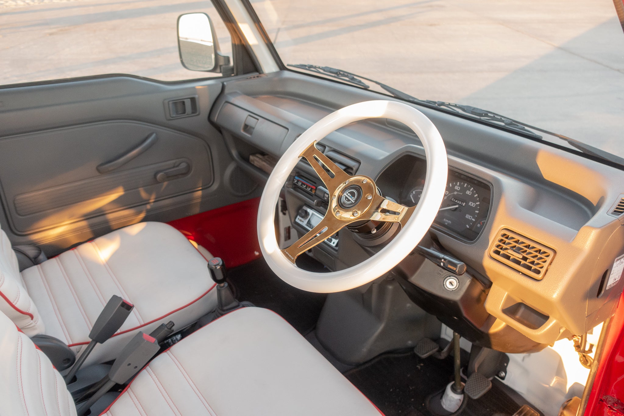 Interior of a Japanese mini truck featuring a white and gold NRG steering wheel, custom beige seats with red trim, and a sleek dashboard design for enhanced driving comfort and style.