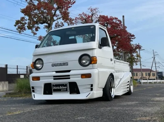 Customized white Suzuki Carry mini truck with lowered suspension and body kit, parked outdoors in a suburban setting with autumn foliage, featuring circular headlights and 'birichino special' license plate.