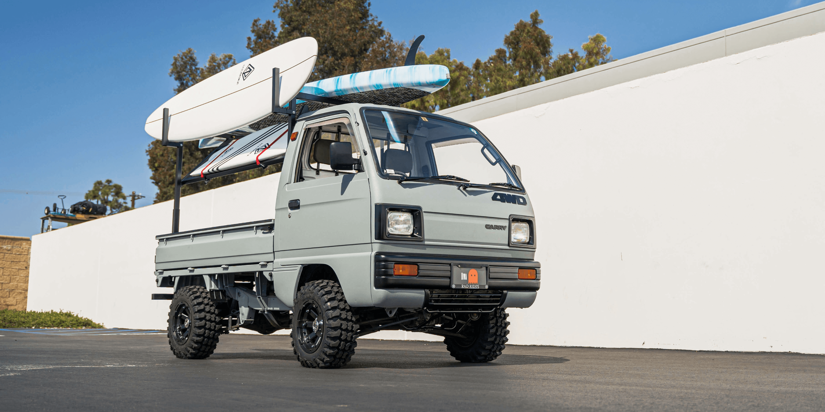 1987 Suzuki Carry DB71T lifted kei truck with off road tires and surfboard rack, light gray Japanese mini truck parked on pavement in front of white wall ready for beach adventure