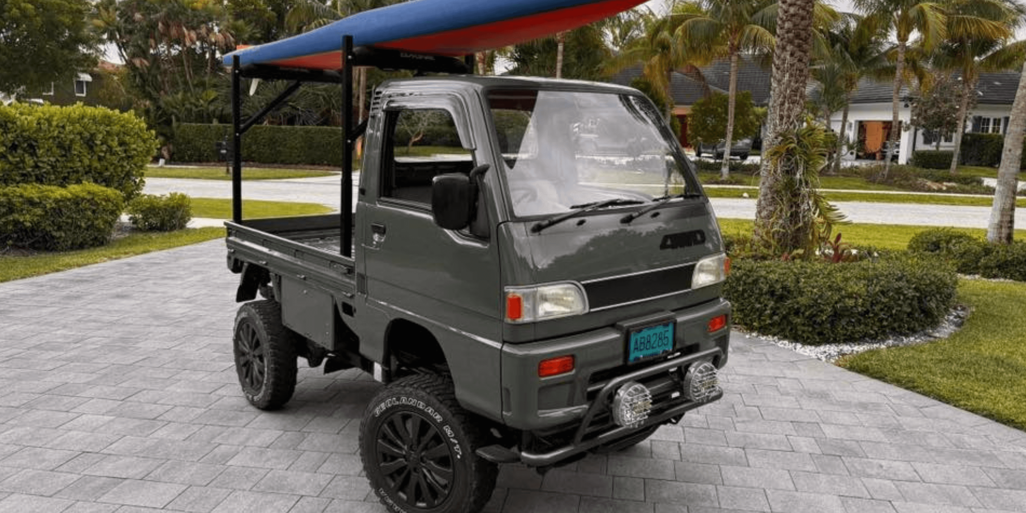 1994 Subaru Sambar KS4 lifted dark green kei truck with black wheels and roof rack carrying a longboard, parked on brick driveway in tropical neighborhood with palm trees