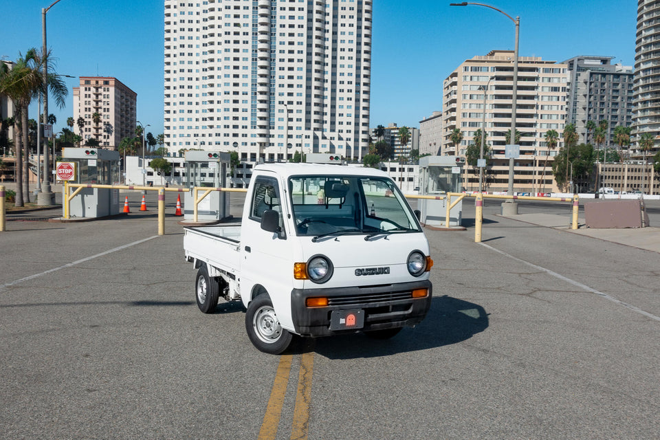 Sleek white Suzuki Carry 9th Generation DC51T mini truck parked in an urban parking lot, showcasing its compact design and versatility in a cityscape backdrop, perfect for efficient urban transportation and cargo handling.