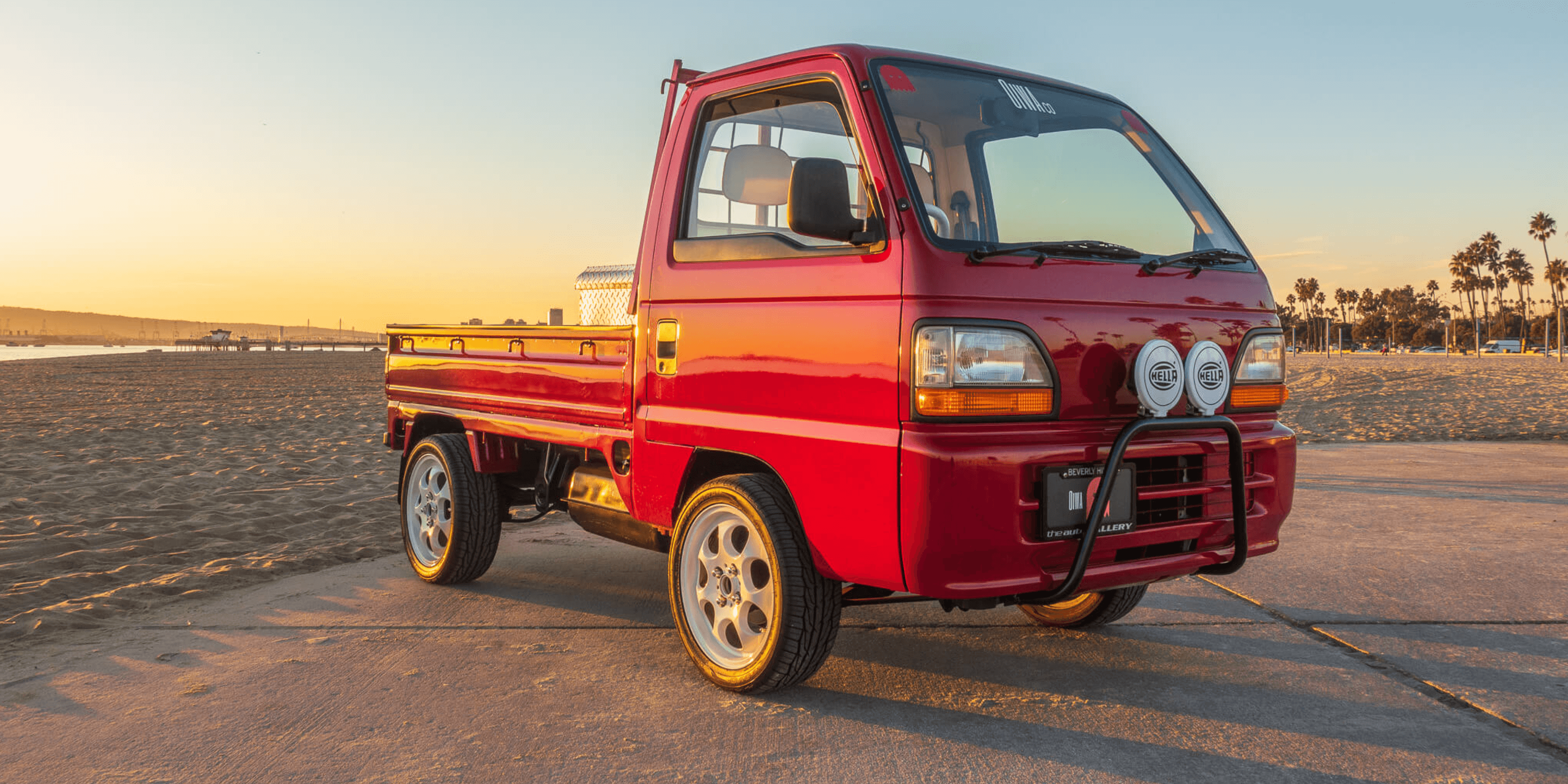 1995 Honda Acty HA4 red kei truck with bull bar and triple rally lights parked on beachside pavement at sunset, classic Japanese mini truck front three quarter view