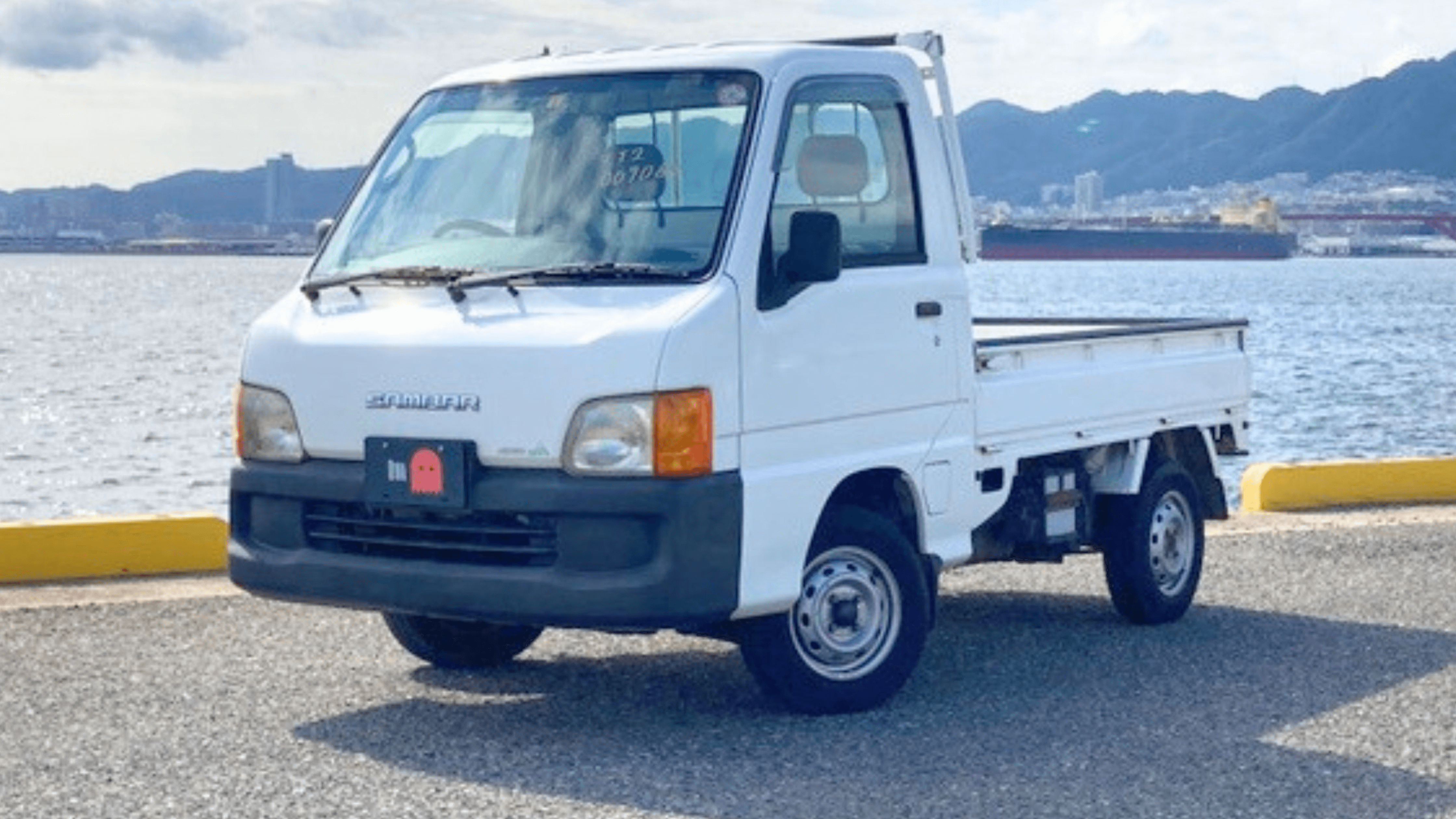 2000 Subaru Sambar TT2 white kei truck parked on waterfront road with mountains and harbor in background front three quarter view showcasing compact Japanese mini truck styling