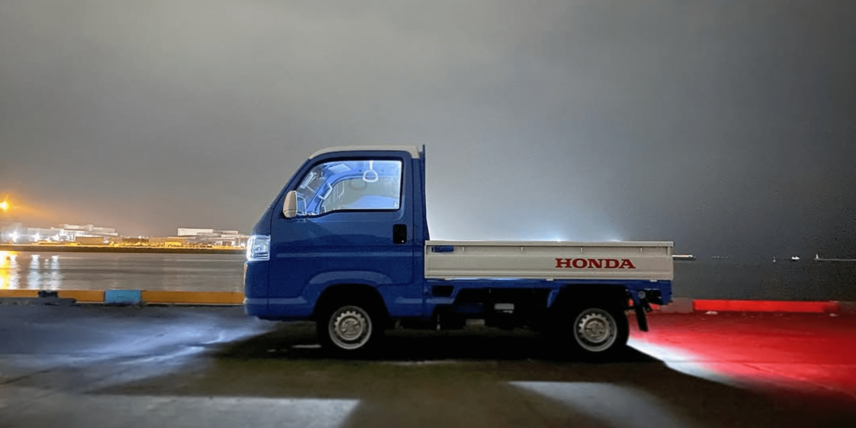 2010 Honda Acty HA9 blue kei truck side profile parked at harbor at night with city lights reflecting on water and red tail lights glowing on wet pavement