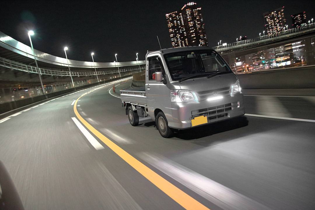 2010 Daihatsu Hijet S211P kei truck in silver with LED daytime running light bar, stock steel wheels, and lowered ride height cruising on a lit city highway at night under urban overpasses – modern Japanese mini truck.