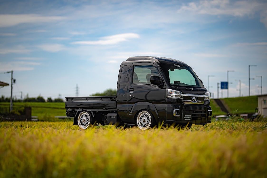 2017 Daihatsu Hijet S510P kei truck in glossy black with lowered suspension, chrome mesh deep-dish alloy wheels, and custom body kit parked in a golden rice paddy field – modern Japanese mini truck styling.