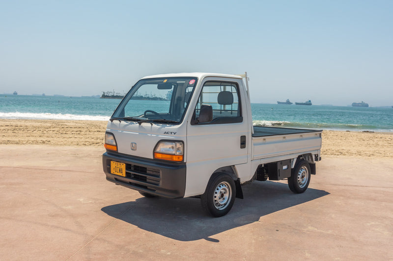Pristine white 1995 Honda Acty HA3 Kei Truck with clear headlights and original Oiwa yellow license plate, parked beachfront, highlighting its compact size, practicality, and classic design, featured on Oiwa Garage’s dedicated Kei truck information page.