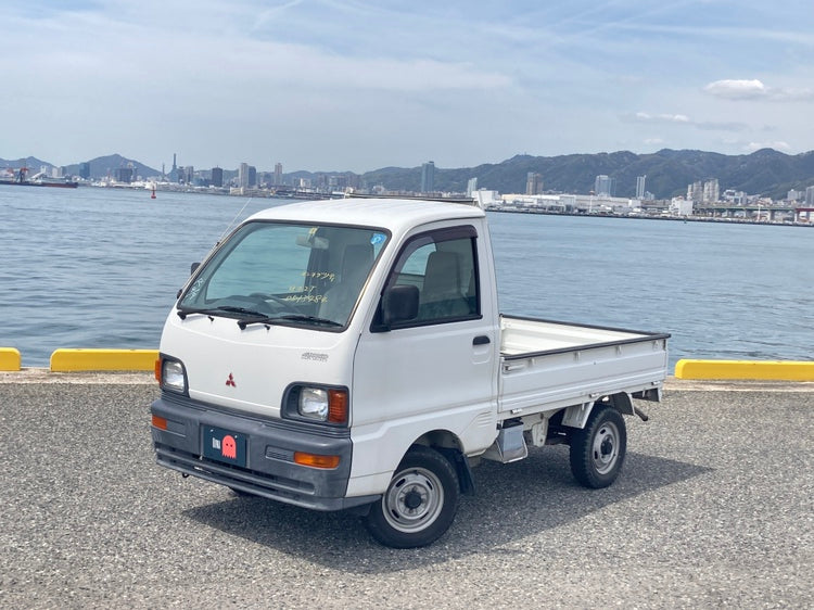 White Mitsubishi Minicab U42T kei truck flatbed parked by a Japanese harbor with city skyline and mountain backdrop.