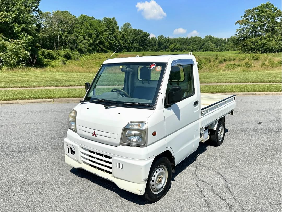1999 Mitsubishi Minicab U62T white kei truck with open flatbed parked on asphalt beside a green field—a compact Japanese mini truck designed for versatile light-duty hauling and deliveries