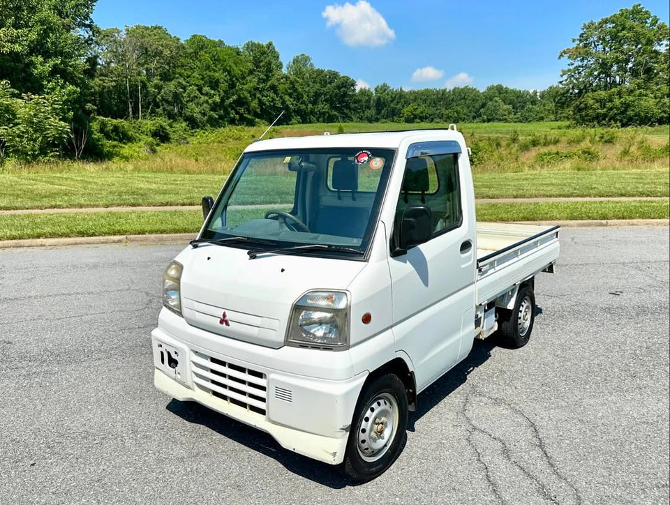 1999 Mitsubishi Minicab U62T white kei truck with open flatbed parked on asphalt beside a green field—a compact Japanese mini truck designed for versatile light-duty hauling and deliveries