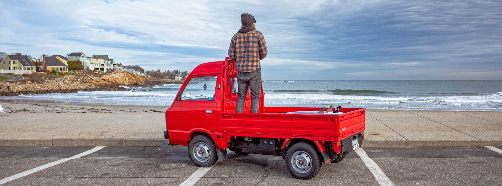 Vibrant red Subaru Sambar KT2 Kei truck parked by a scenic beach, featuring a man standing in the bed overlooking the ocean, symbolizing freedom and the truck's capability for recreational and commercial activities in picturesque locations.